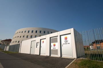 Budova Centrální depozitář UPM se zaoblenými okny vedle moderní trafostanice s nápisy na dveřích, oplocený prostor pod modrou oblohou.