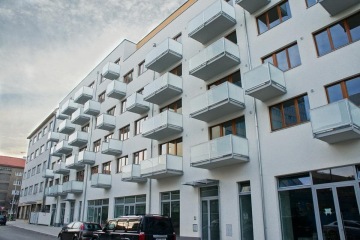 Moderní bytový dům s balkóny z mléčného skla a zaparkovanými auty před vchodem.