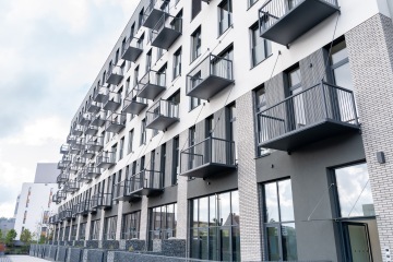 Moderní bytový dům s řadou balkónů a velkými okny, jasně modrá obloha v pozadí.