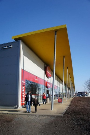 Obchod Hervis s lidmi odcházejícími ze vchodu, modrá obloha a žlutý přístřešek.