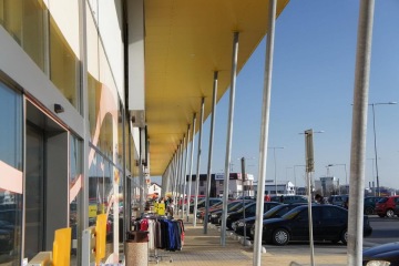 Nákupní středisko s dlouhým skleněným průčelím, parkovištěm plným aut a vystaveným oblečením před obchodem.