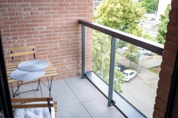 Malý balkon s dřevěným stolem a židlemi, z cihelné zdi, výhledem na ulici s parkujícími auty a zelení.