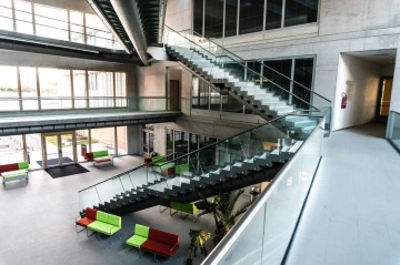 Moderní vnitřní prostor Jihočeská Univerzita s kovovým schodištěm, skleněnými zábradlími a světlem osvětlenými pohovkami v zelené a červené barvě.