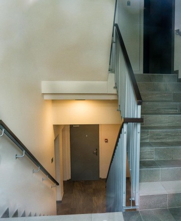 Schodiště v budově BD Nad Modřanskou s moderním zábradlím a osvětlením, vedoucí ke dveřím v přízemí.