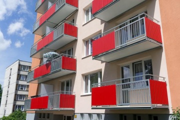 Bytový dům s červenými závěsnými balkony a modrým slunečníkem na jednom z nich pod modrou oblohou.