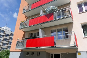 Panelový dům s červenými závěsnými balkony a modrou oblohou v pozadí.