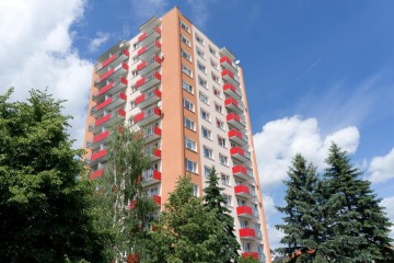 Panelová budova se světlou fasádou a červenými závěsnými balkony, obklopená zelení proti modré obloze.