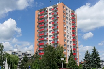 Vysoký panelový dům s červenými závěsnými balkony pod modrou oblohou, obklopený stromy.