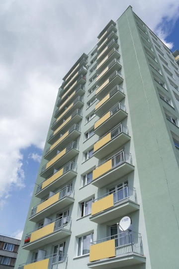 Vysoký panelový dům se žlutými závěsnými balkony proti oblačné obloze.