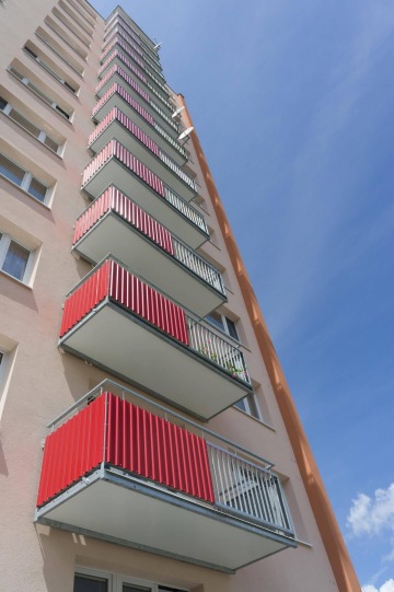 Panelový dům s červenými závěsnými balkony na pozadí modré oblohy.