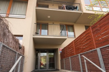 Vchod do moderního bytového domu s balkony a kovovým oplocením.