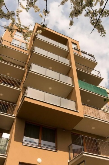 Moderní bytový dům s balkony pod modrou oblohou, obklopený kvetoucími stromy.