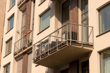 Balkon městské budovy X Loft s kovovým zábradlím, kolo a nábytek, květináče na vedlejším balkonu.
