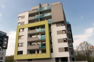 Moderní bytový dům s balkony a zeleným rámem proti modré obloze.