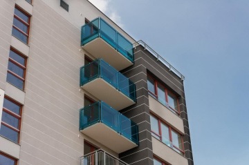 Moderní bytový dům s modrými balkony a červenými okenními rámy pod modrou oblohou.