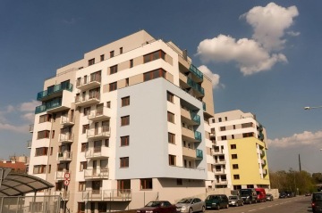 Moderní bytové domy s balkony a zaparkovanými auty pod modrou oblohou.