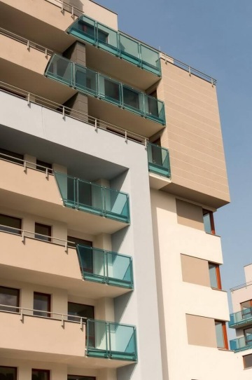 Moderní bytový dům s balkony se skleněnými zábradlími a modrými rámy proti modré obloze.