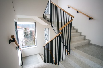 Moderní schodiště s kovovým zábradlím a dřevěnými madly vedoucími kolem velkých oken s výhledem na venkovní krajinu.
