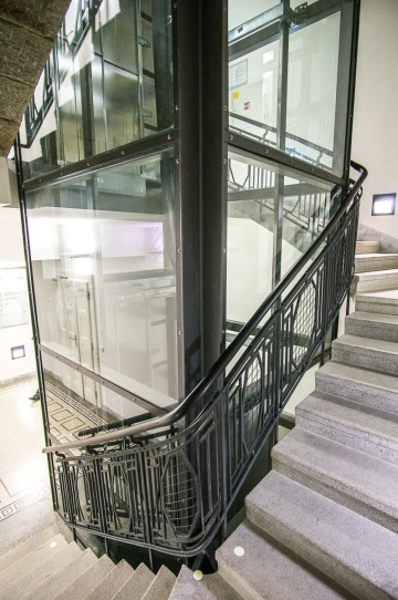 Prosklená výtahová šachta obklopená točitým schodištěm s kovovým zábradlím.