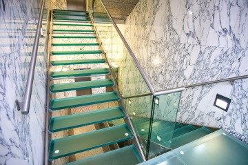 Skleněné schodiště s kovovým zábradlím v interiéru s mramorovými stěnami.