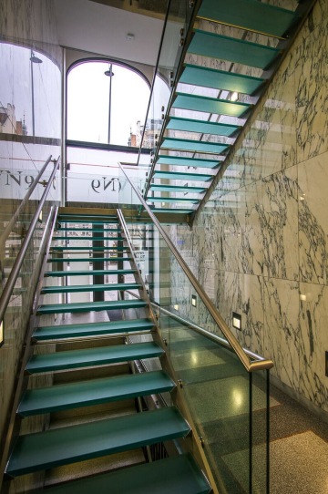 Skleněné schodiště s kovovým zábradlím, mramorové stěny, velké okno.