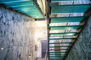 Moderní schodiště s prosklenými schody a mramorovými stěnami vedle výtahů.