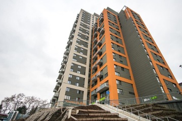 Vysoká moderní budova s oranžovými a šedými fasádami, schodiště a chodník v popředí.