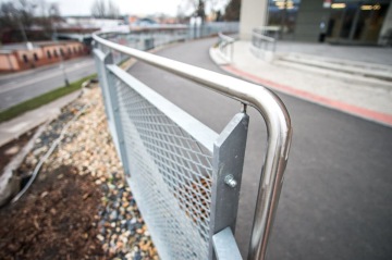 Moderní kovové zábradlí nad chodníkem s výhledem na ulici a budovu v pozadí.