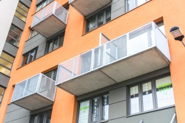 Moderní bytový dům s oranžovou fasádou a balkony z kovového pletiva, v přízemí jsou komerční prostory s nápisy na oknech.