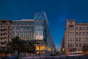 Moderní skleněná budova s vlnitým designem fasády za soumraku, obklopená historickými budovami.