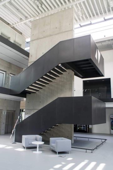 Moderní interiér Laserové centrum ELI s černým schodištěm a šedými křesly u kulatého stolku, betonová stěna, prosvětlený prostor.