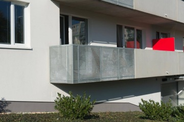 Balkon s perforovaným kovovým zábradlím na moderním bytovém domě BD Zahrady Opatov, červený panel, zelené keře vpředu.
