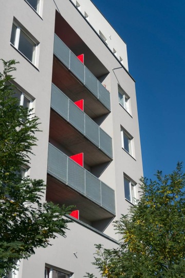 Moderní bytový dům BD Zahrady Opatov s balkony, šedou fasádou a červenými panely, obklopený stromy na pozadí modré oblohy.