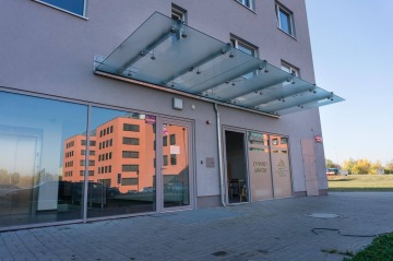 Vstup do moderní budovy BD Zahrady Opatov s prosklenými dveřmi a skleněným přístřeškem, v pozadí oranžová budova a travnatý prostor.