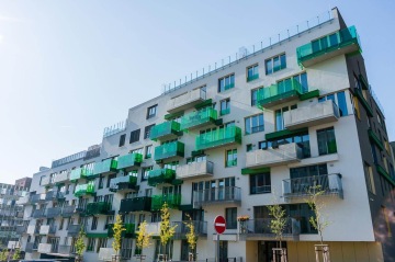 Moderní bytový dům Tesla Vršovice se zelenými balkony a mladými stromy v popředí pod modrou oblohou.