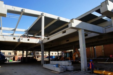 Konstrukce rozestavěné budovy s pracovníkem v helmě, bagrem a žebříkem pod modrou oblohou.