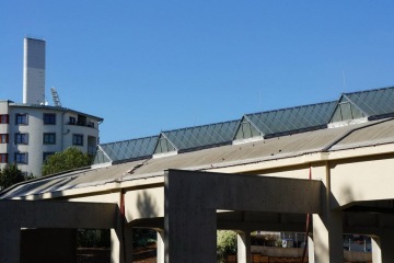 Šedá budova s moderní střechou, vedle níž je vícepatrový dům, pod modrou oblohou.