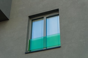 Zeleně zabarvené okno s žaluziemi na šedé omítkové zdi budovy Tesla Vršovice.