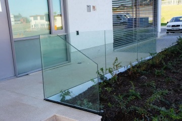 Vnější skleněné zábradlí v moderním exteriéru u budovy, vedle zeleného záhonu a zaparkovaného auta.