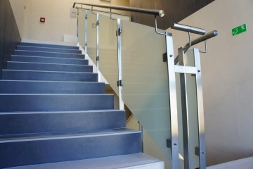 Moderní vnitřní schodiště s kovovým zábradlím a skleněnou výplní, v pozadí je vidět únikové značení.