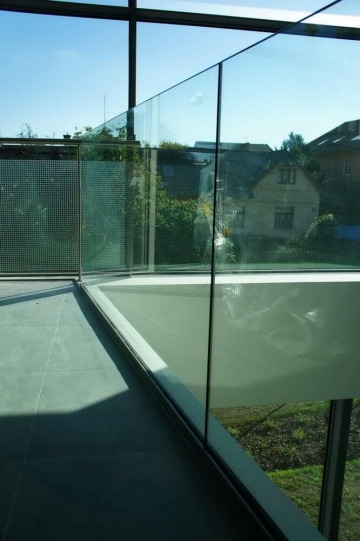 Balkon s moderním vnějším skleněným zábradlím a výhledem na stromy a domy za slunečného dne.