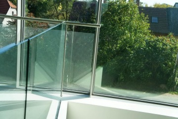 Vnější moderní prosklené zábradlí balkónu s výhledem na zahradu a střechy domů.