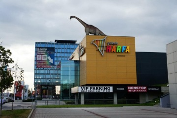 Barevná fasáda Galerie Harfa s velkou sochou dinosaura na střeše za oblačného počasí.