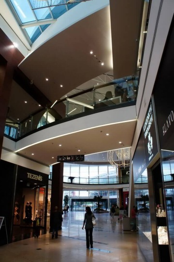 Moderní obchodní centrum Galerie Harfa s více úrovněmi, prosklené stěny a osvětlení. Lidé procházejí kolem obchodu s názvem Tezenis.