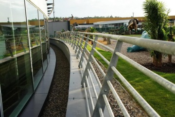 Zábradlí vedoucí podél prosklené budovy Galerie Harfa s výhledem na trávník a umělé dinosaury v dálce.