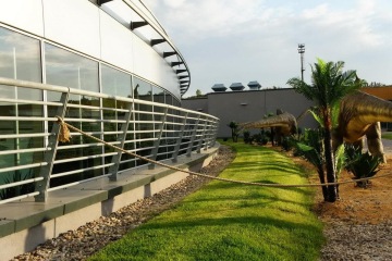 Moderní budova Galerie Harfa s prosklenými stěnami, vedle ní modely dinosaurů a tropické rostliny na travnatém povrchu.