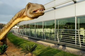 Dinosaurus stojící u moderní budovy Galerie Harfa s prosklenou fasádou a zeleným trávníkem.