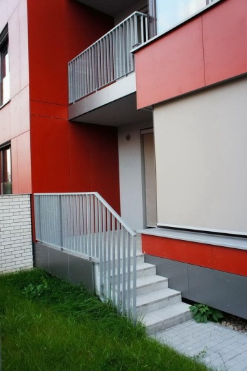 Moderní bytový dům s červenou fasádou, balkonem a schodištěm s kovovým vnějším zábradlím.