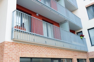 Předsazený balkon s vnějším kovovým zábradlím, květináčem a cihlami na fasádě budovy.