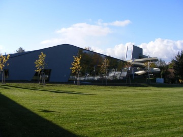 Moderní budova se zelenou střechou, prosklenými stěnami a skluzavkami, obklopená trávníkem a stromy na slunečném dni.